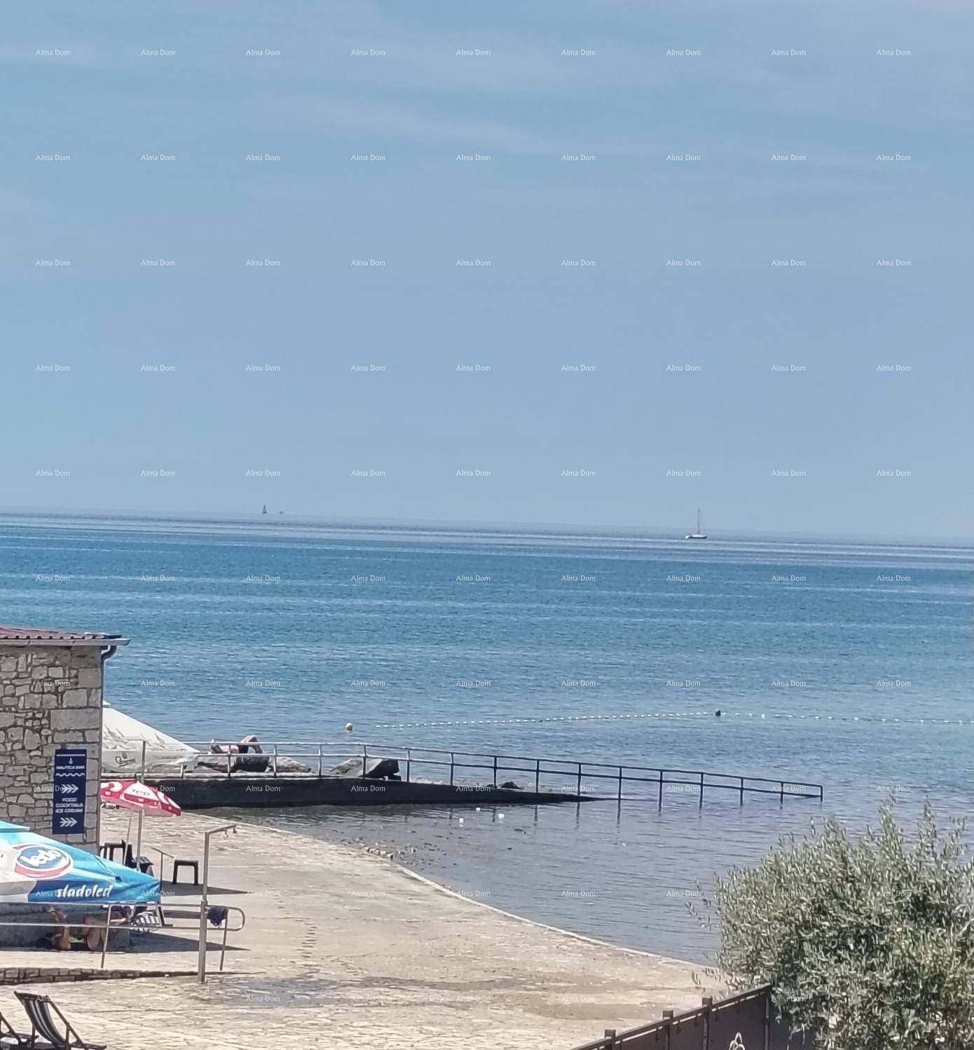 Panoramablick auf das ruhige Meer mit einem Sandstrand, einem kleinen Steg und einem Segelboot am Horizont.
