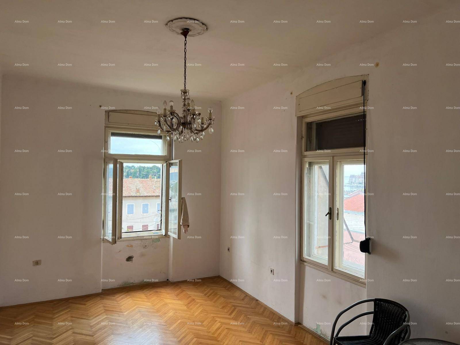 Geräumiges Zimmer mit Fischgrätparkett, zwei Fenstern und einem eleganten Kronleuchter, bereit zur Renovierung.