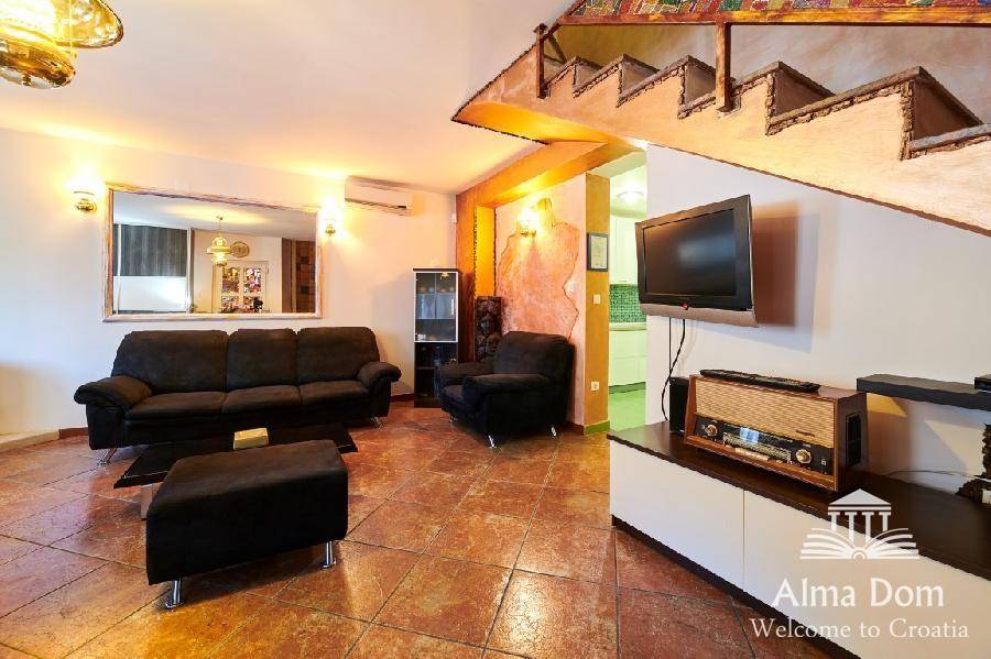 Wohnbereich mit Fernseher, Sofas und einer kunstvoll gestalteten Treppe im Hintergrund.