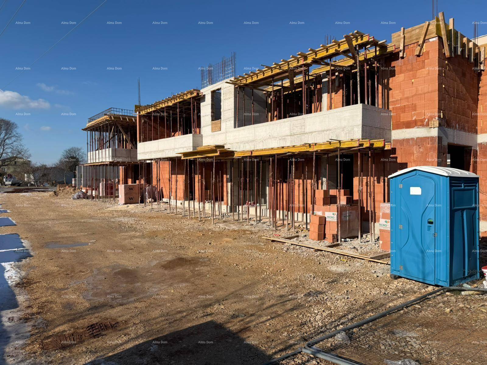 Baustelle eines Neubaus mit sichtbaren Ziegelwänden, Schalung und einem mobilen WC unter blauem Himmel.
