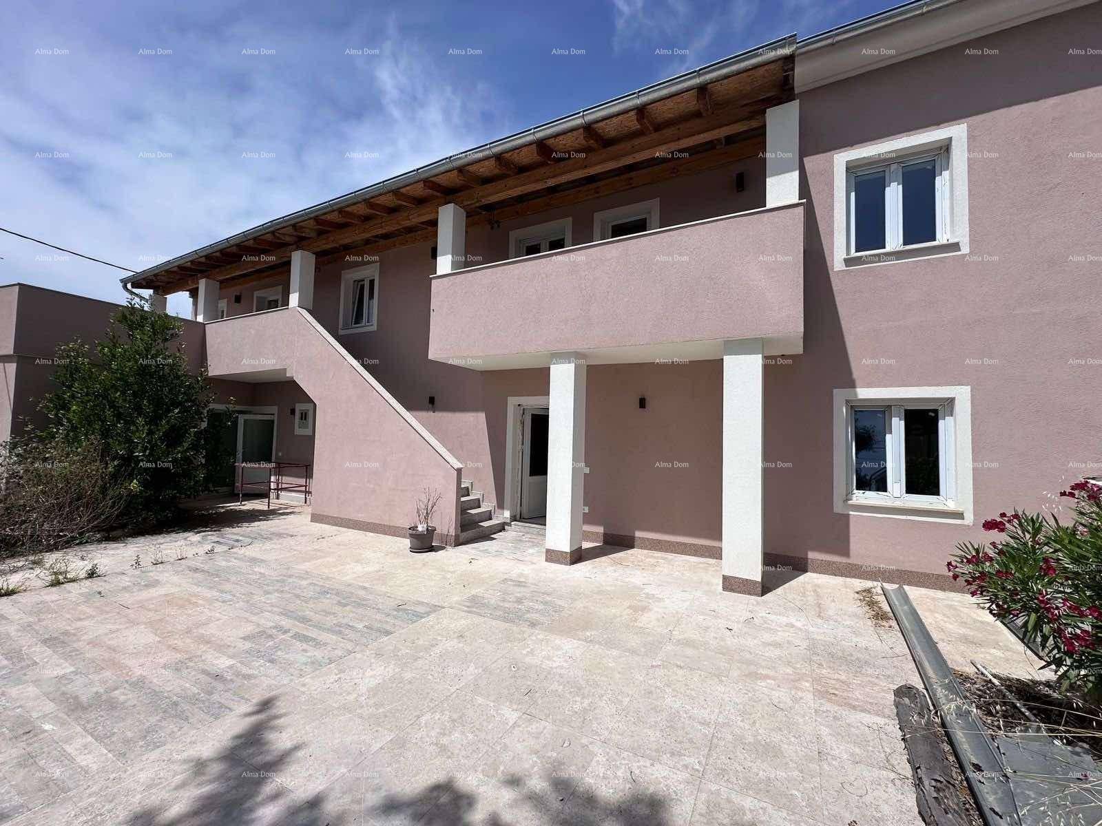 Frontansicht des Gebäudes mit Balkonen, Außentreppe und gepflastertem Vorplatz in der Sonne.