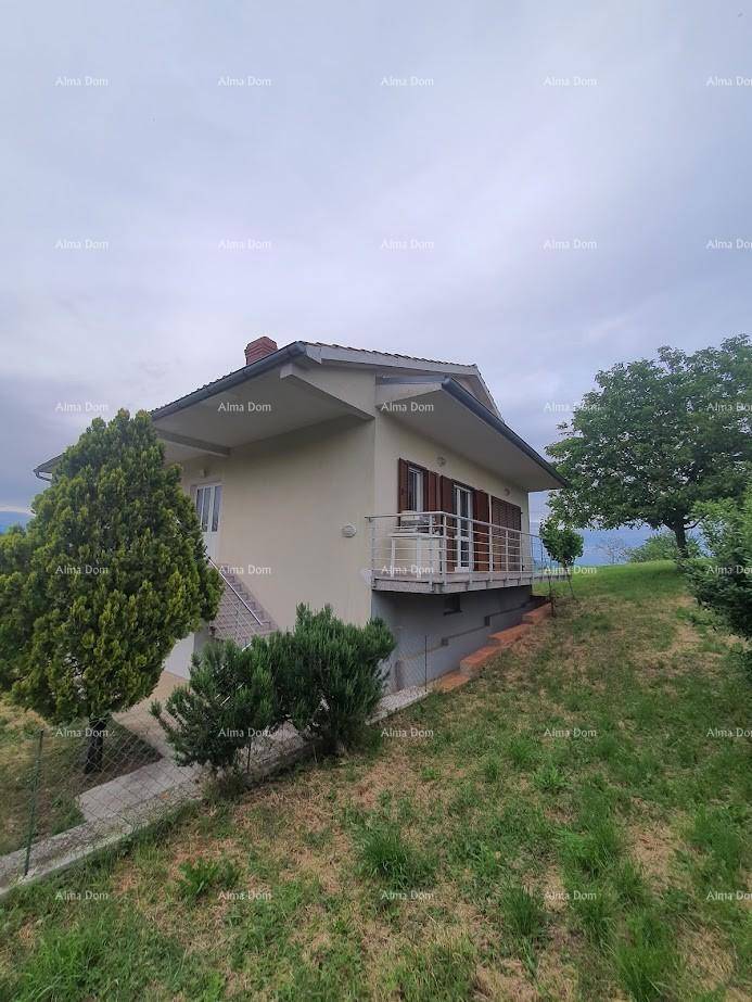 Seitenansicht des Hauses mit Balkon, umgeben von Bäumen und Sträuchern unter bewölktem Himmel.