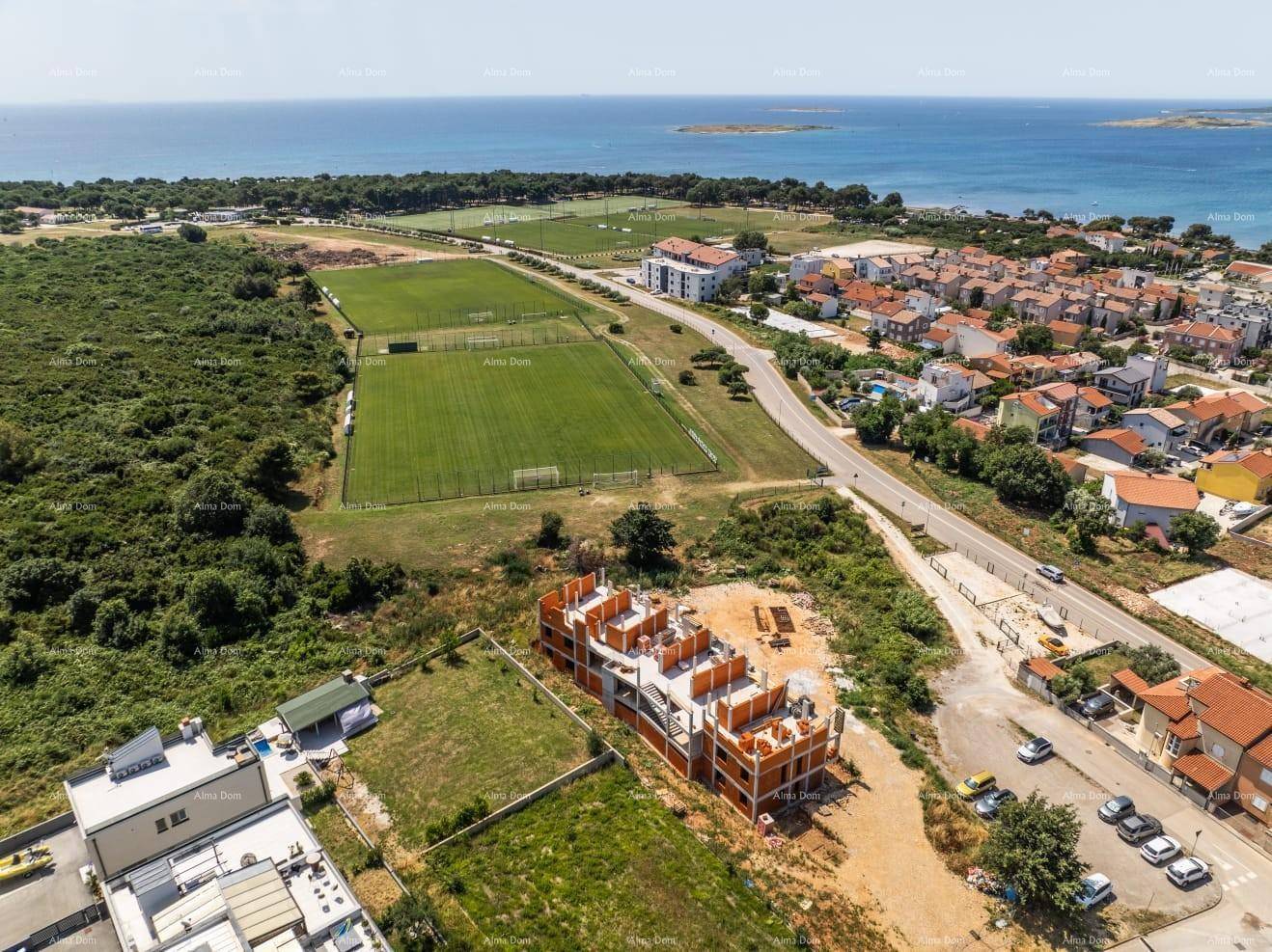 Luftaufnahme der Baustelle mit Sportplätzen, Straße und dem Meer im Hintergrund.