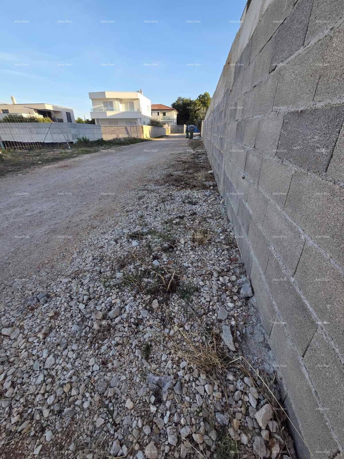 Schotterweg neben einer Betonsteinmauer mit modernen Häusern im Hintergrund unter blauem Himmel.