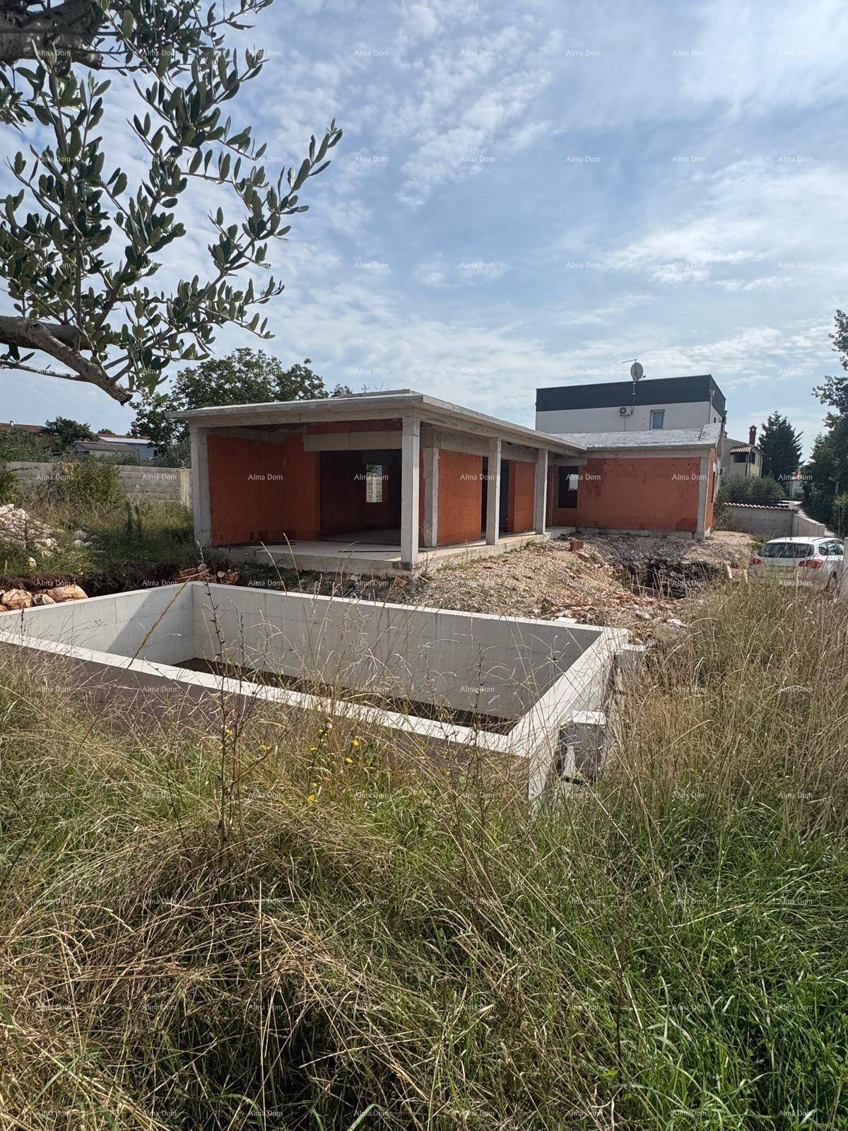 Baustelle mit Rohbau eines Hauses, Poolfundament und viel Grün in der Umgebung unter bewölktem Himmel.