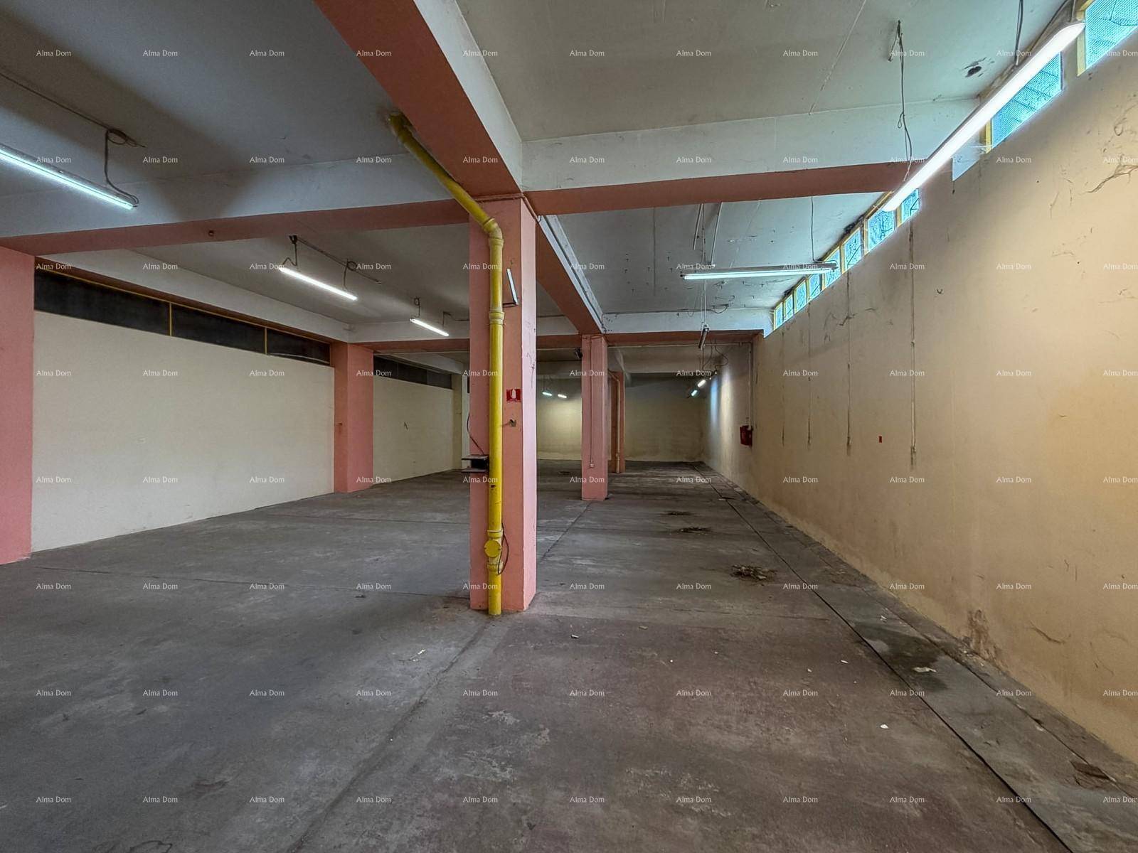 Weiter Blick in den leeren Lagerraum mit Betonboden, rosa Stützpfeilern und gelben Rohren an der Decke.