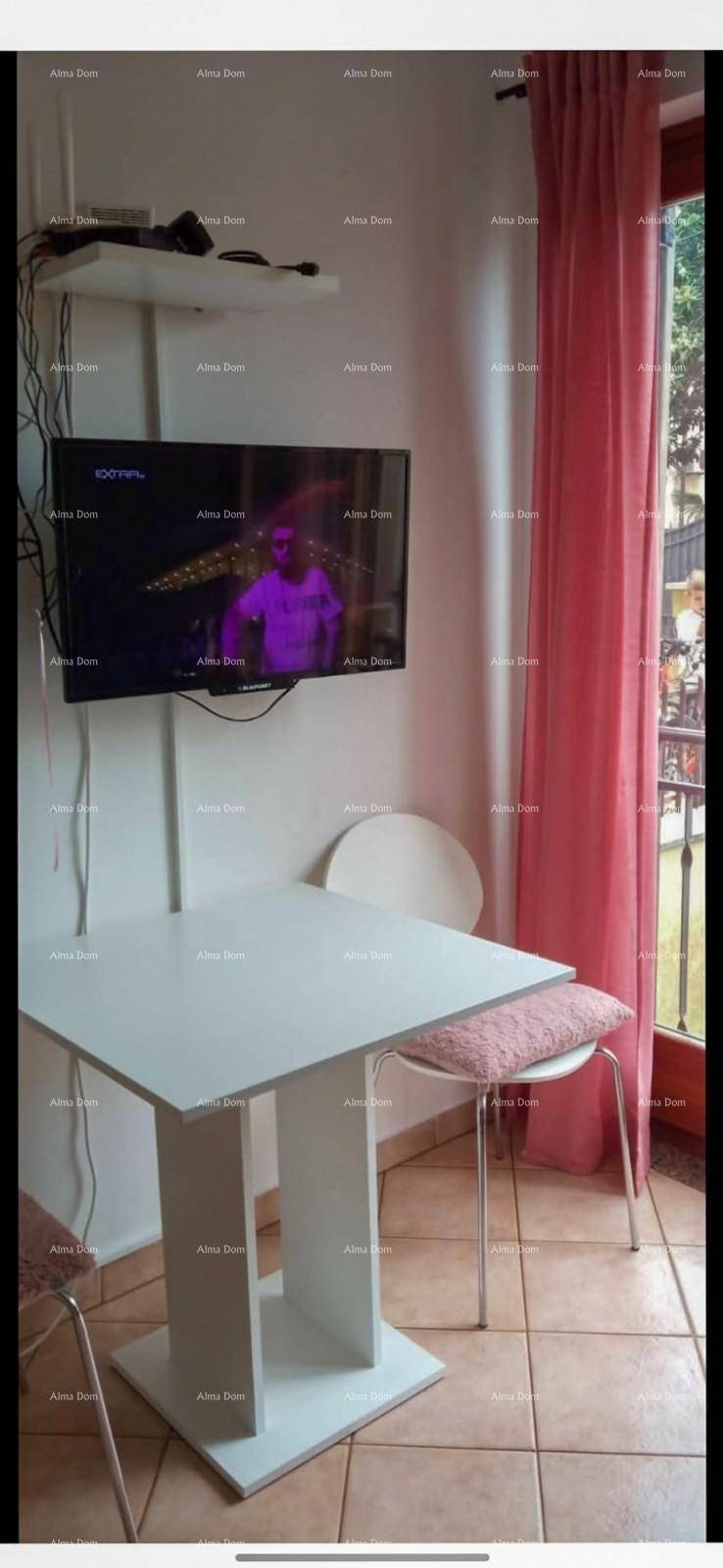 Kleiner Esstisch mit Stühlen, Wandfernseher und Fenster mit rosa Vorhängen in einem Wohnbereich.