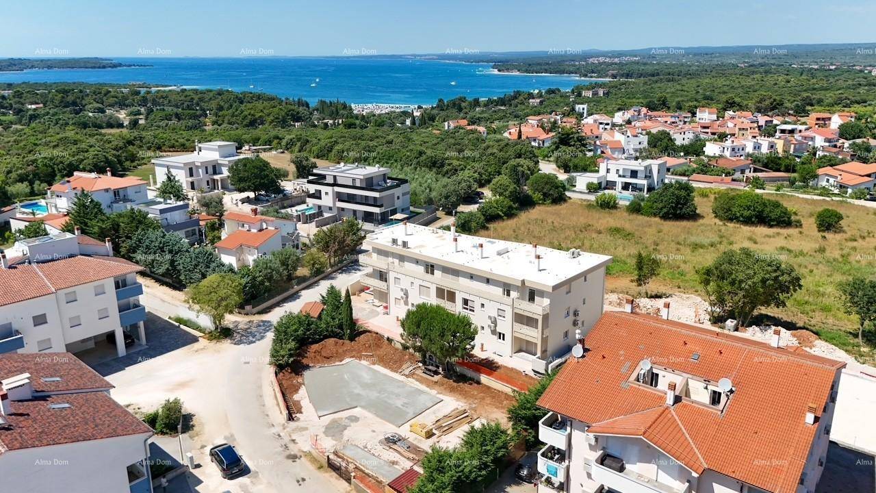 Luftaufnahme einer Küstensiedlung mit Wohngebäuden, viel Grün und dem Meer im Hintergrund unter blauem Himmel.