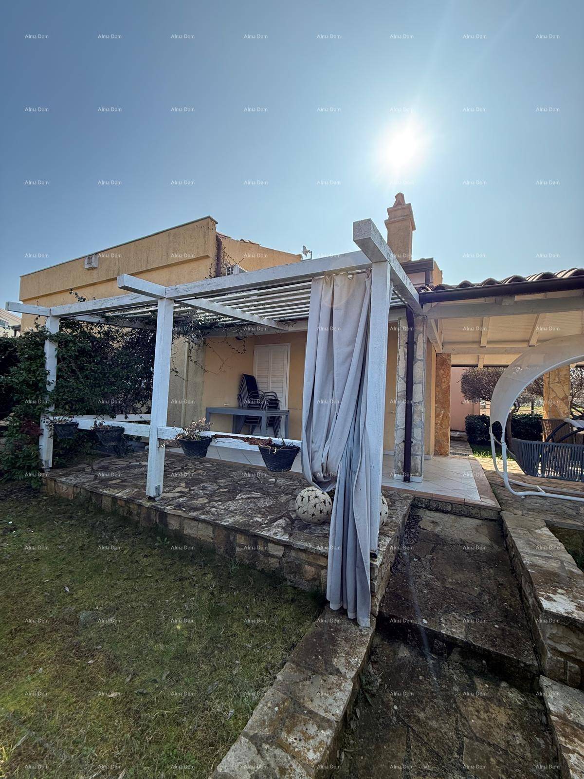 Sonniger Außenbereich mit Pergola und Vorhängen, angrenzend an das Gebäude mit Steinfassade.