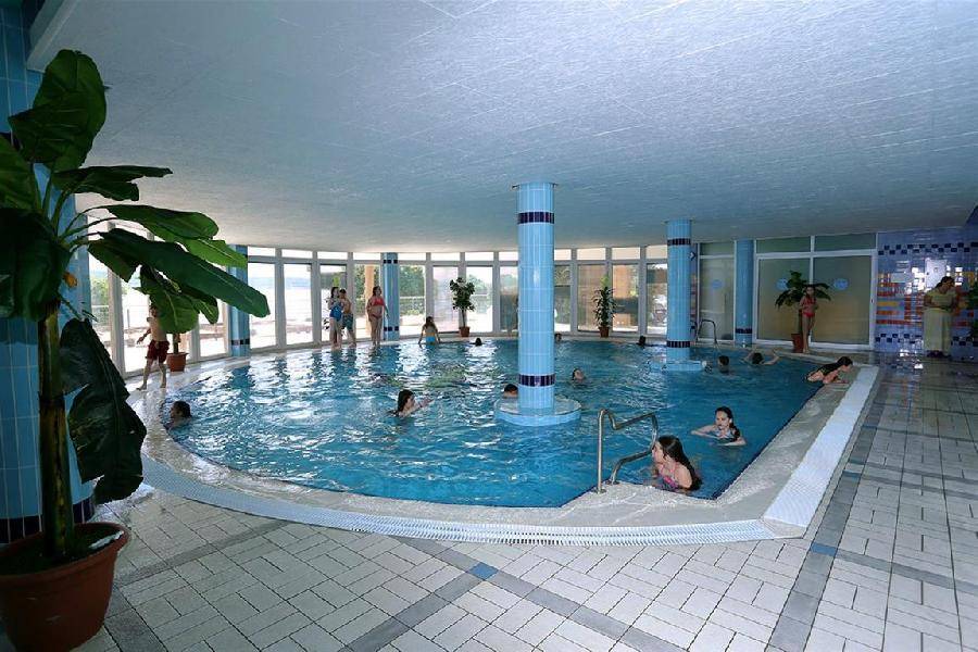 Großes Hallenbad mit vielen Badegästen, umgeben von großen Fenstern und hellen Fliesen.