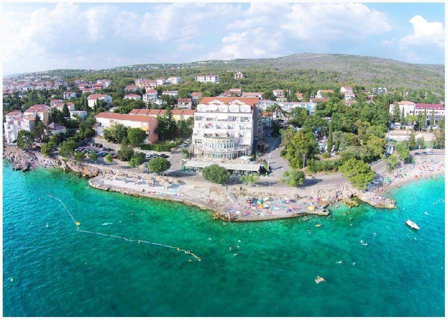 Luftaufnahme eines Hotels mit Strandabschnitt, umgeben von Wohngebäuden und üppiger Vegetation am Meer.