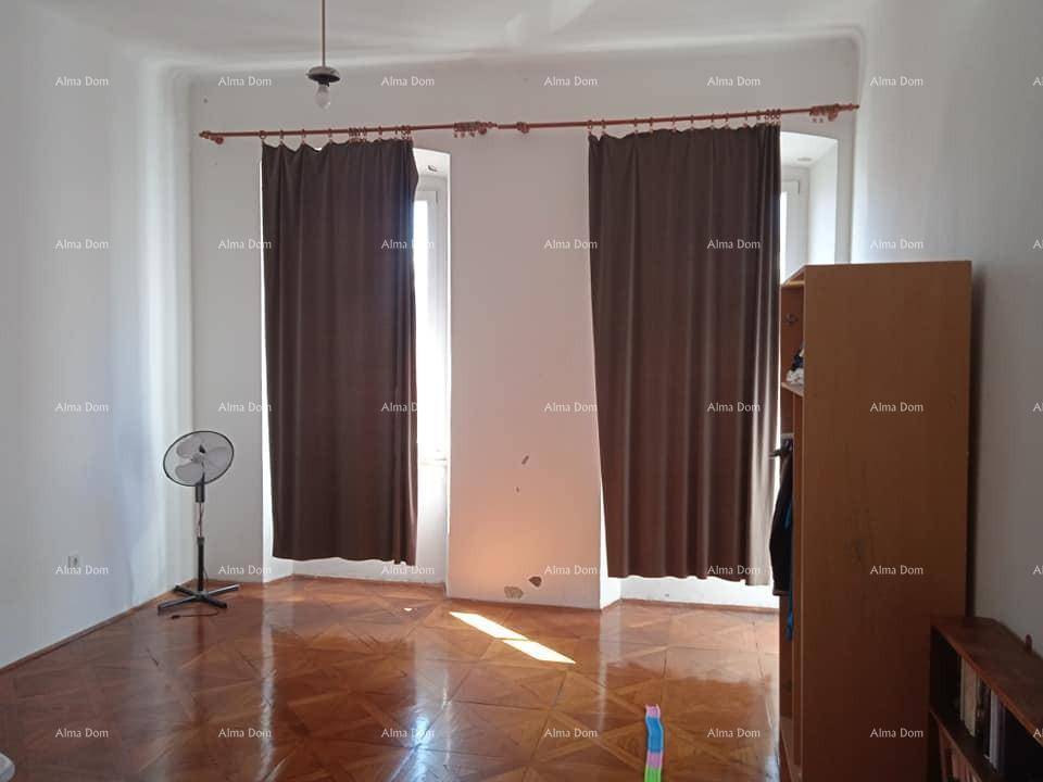 Heller Raum mit Parkettboden, zwei Fenstern mit dunklen Vorhängen und einem stehenden Ventilator.