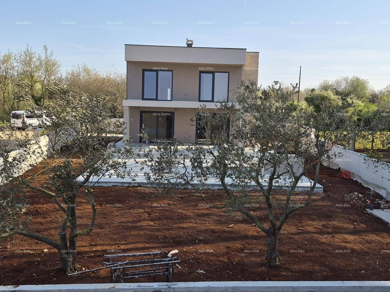Vorderansicht eines modernen Hauses im Bau mit vorbereitetem Gartenbereich und Olivenbäumen.