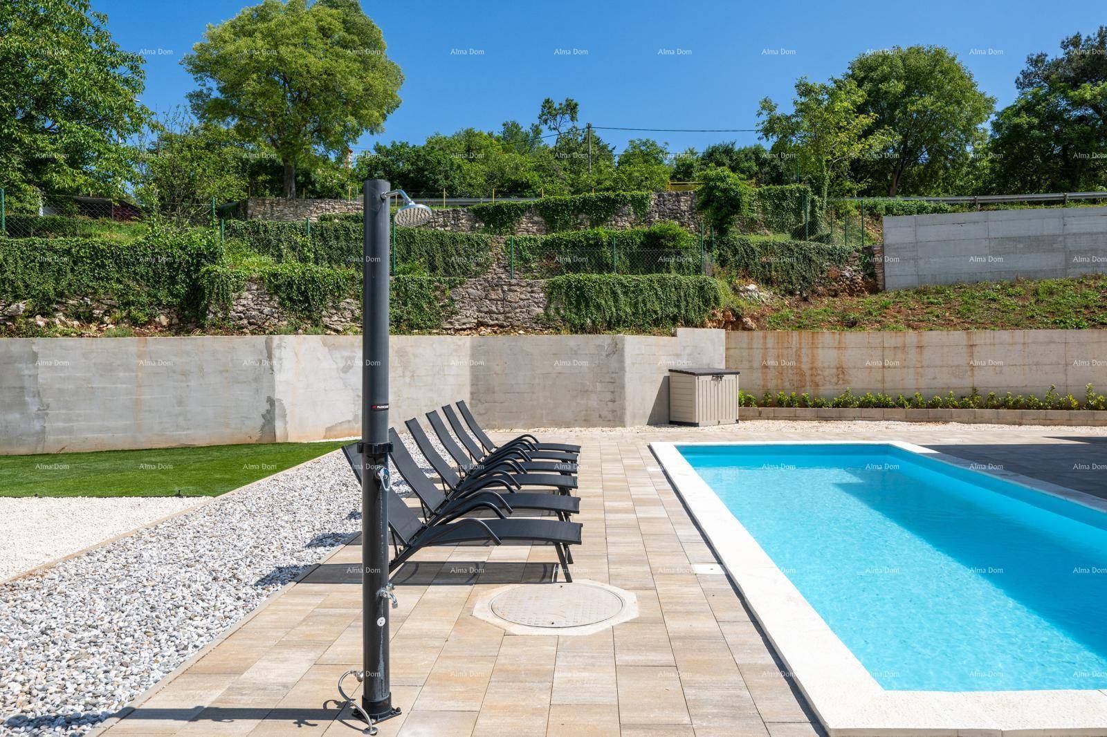 Poolbereich mit Außendusche und mehreren Liegestühlen, umgeben von Grünflächen.
