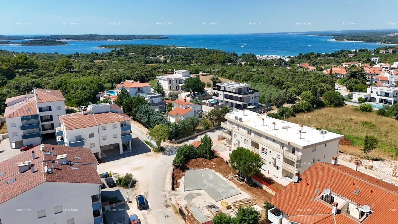 Luftaufnahme einer Küstensiedlung mit Wohngebäuden, viel Grün und dem Meer im Hintergrund unter blauem Himmel.