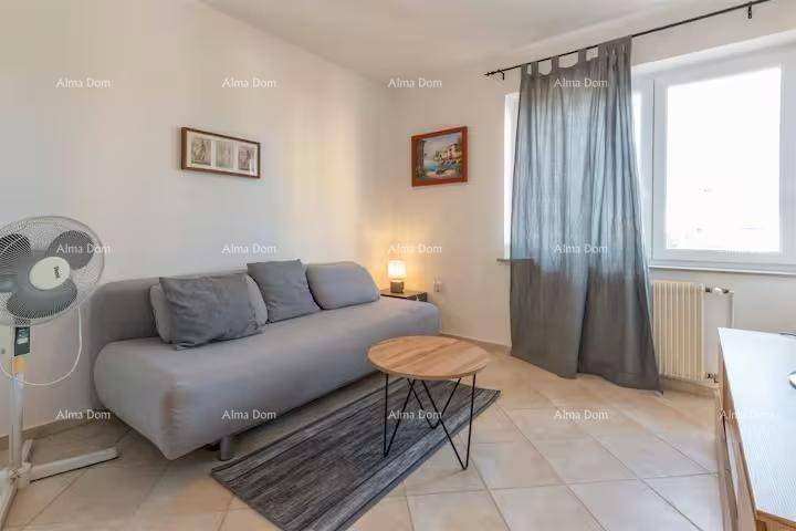 Helles Wohnzimmer mit beigem Fliesenboden, grauem Sofa und Fenster mit Vorhängen.