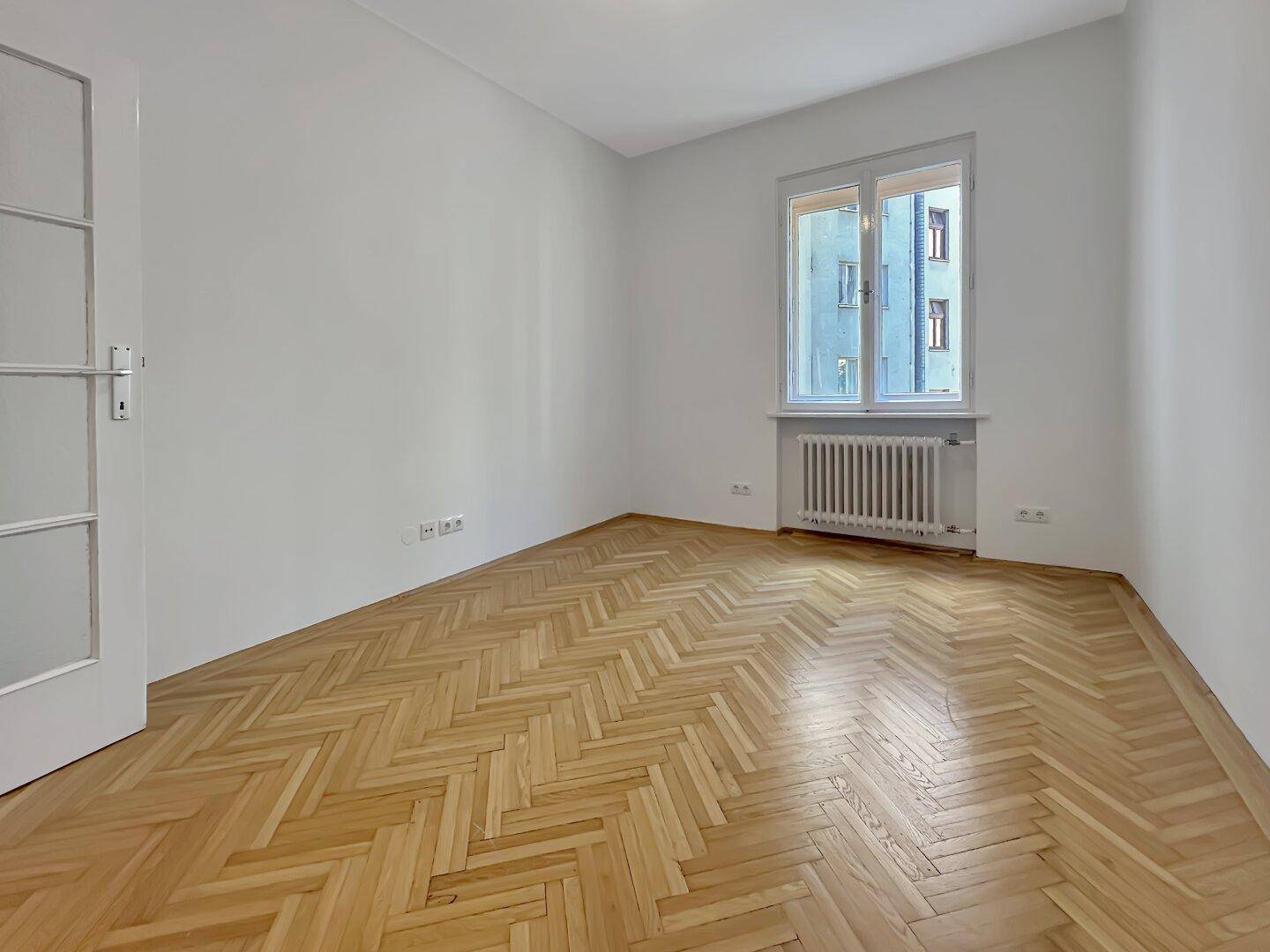 Gemütliches Zimmer mit Fischgrätparkett, weißen Wänden und einem Fenster mit Blick nach draußen.