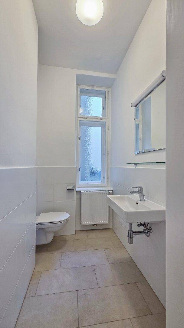 Helles Badezimmer mit Fenster, WC, Waschbecken und weißen Fliesen an Wänden und Boden.