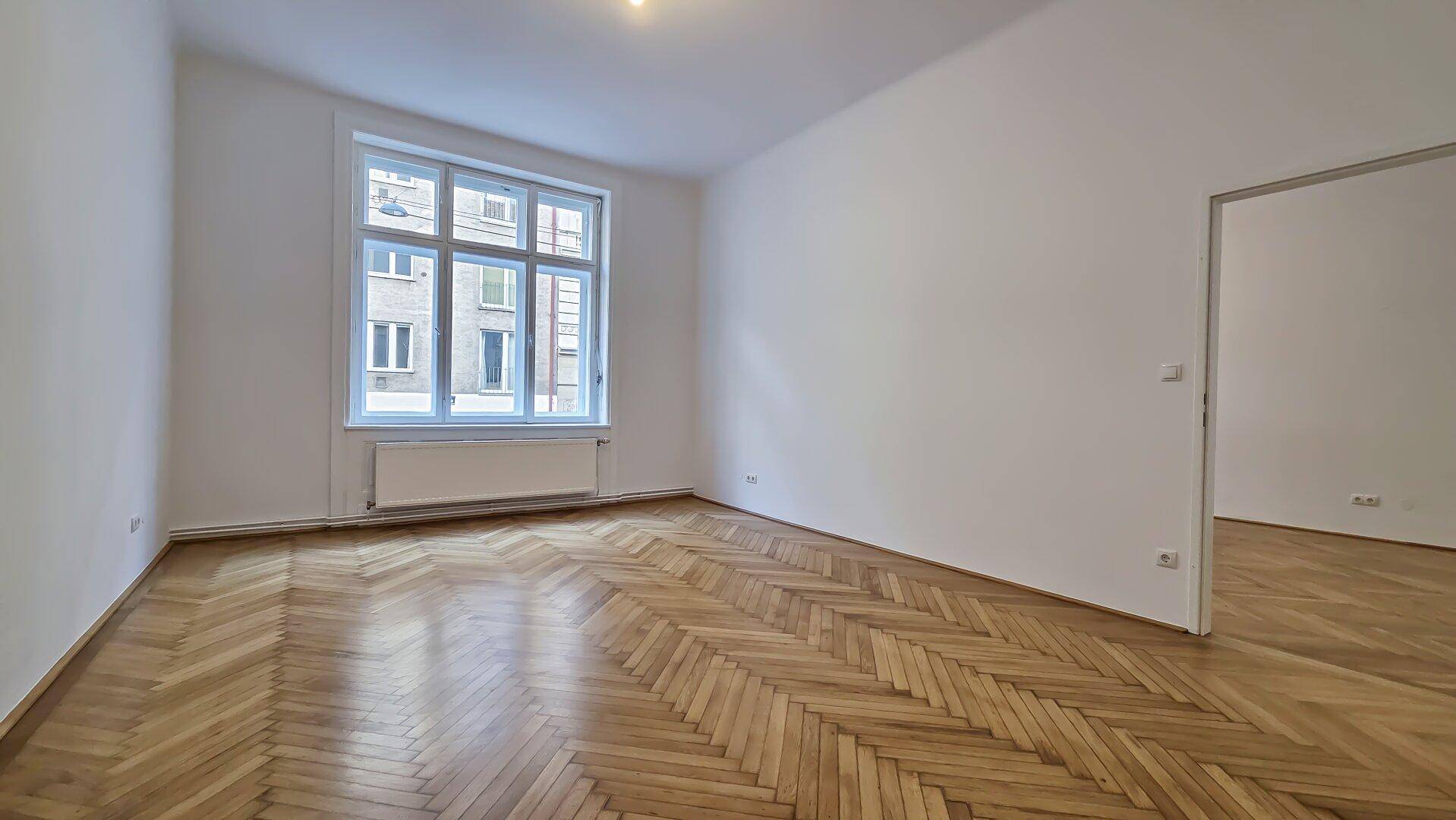 Heller Raum mit elegantem Fischgrätparkett und einem großen Fenster, das den Blick auf die Straße freigibt.