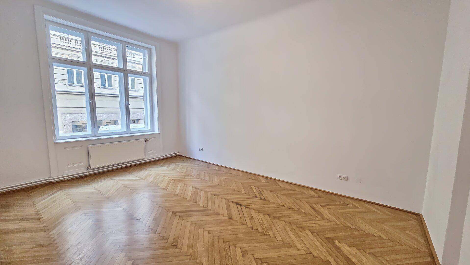 Heller Wohnraum mit klassischem Fischgrätparkett und einem großen Fenster, das den Blick auf die gegenüberliegende Fassade bietet.