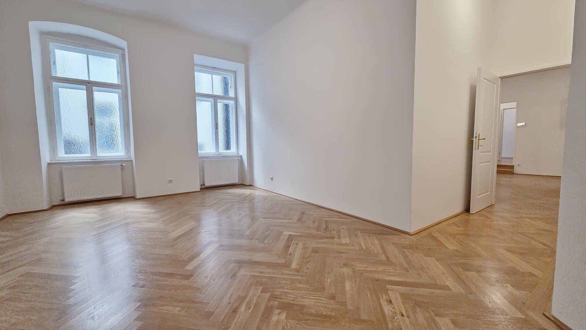 Geräumiges, helles Zimmer mit klassischem Fischgrätparkett und zwei großen Fenstern, die viel Tageslicht hereinlassen.