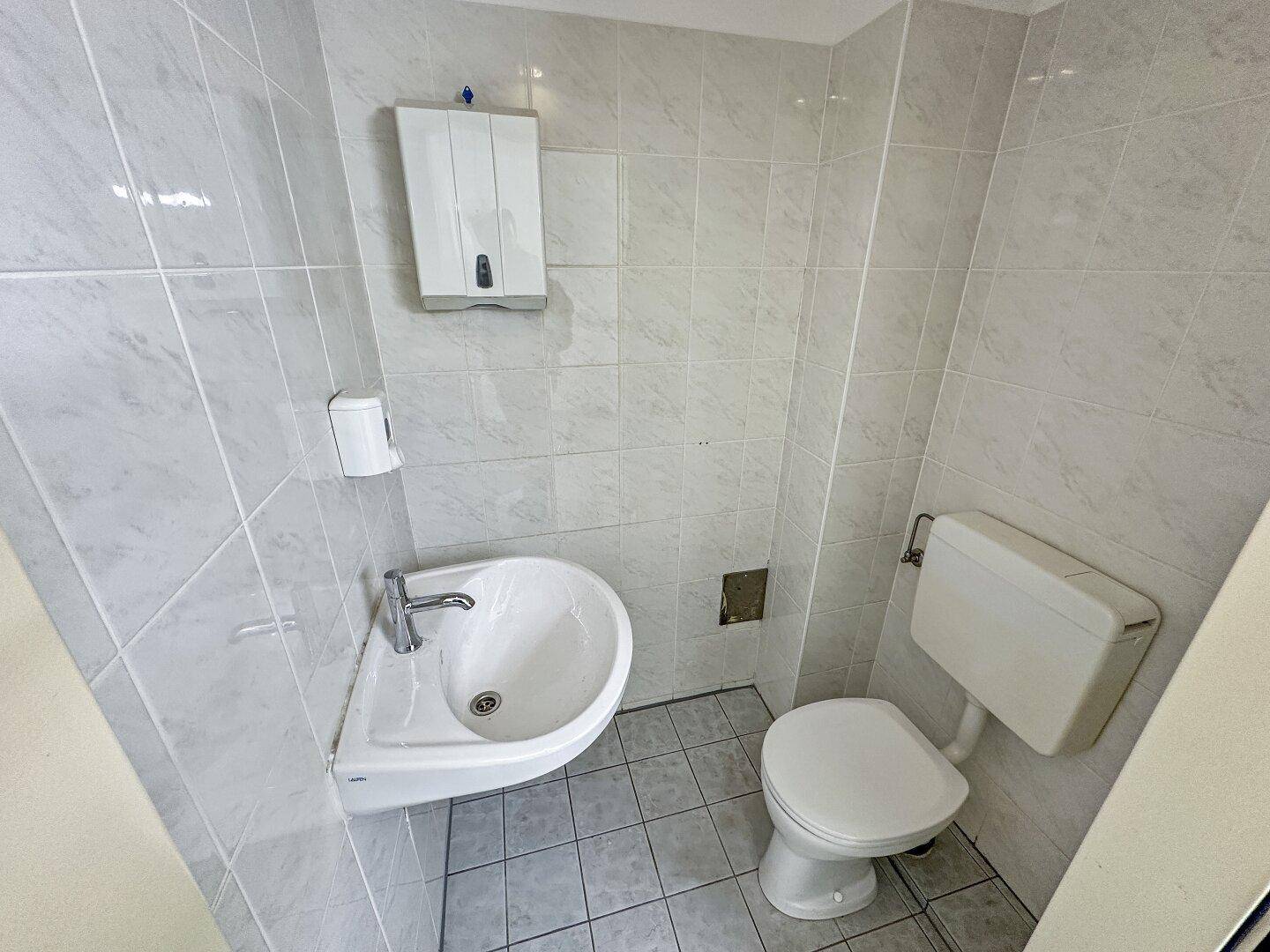 Kleines WC mit Eckwaschbecken, Toilette und hellen Fliesen an Wänden und Boden.
