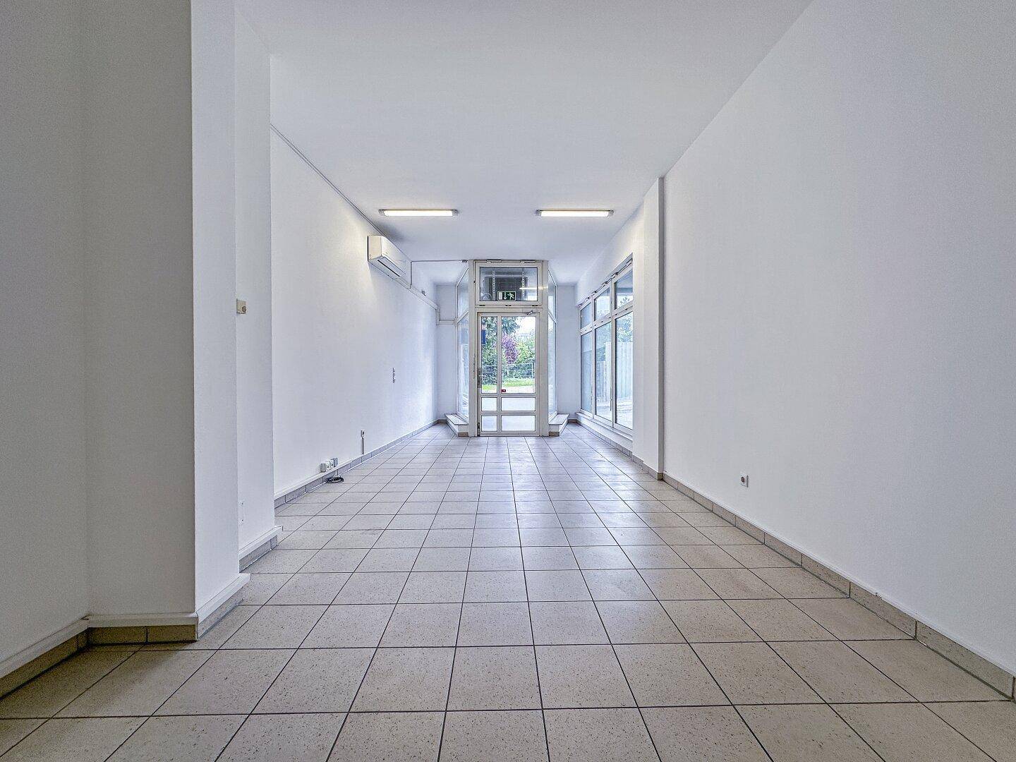 Langer, leerer Gewerberaum mit Fliesenboden, weißen Wänden und viel Tageslicht durch die Glasfront.