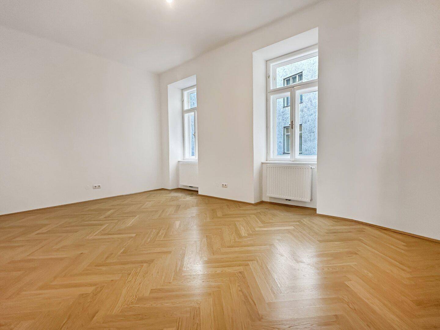 Helles Zimmer mit Fischgrätparkett und zwei hohen Fenstern, die viel Tageslicht hereinlassen.