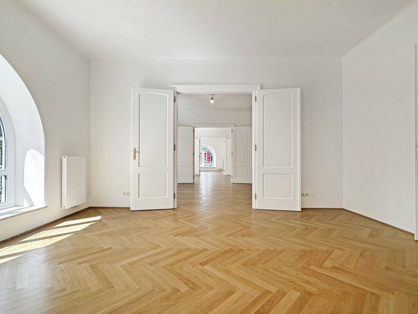 Geräumiges Zimmer mit Fischgrätparkett und hohen weißen Türen, die zu weiteren Räumen führen.