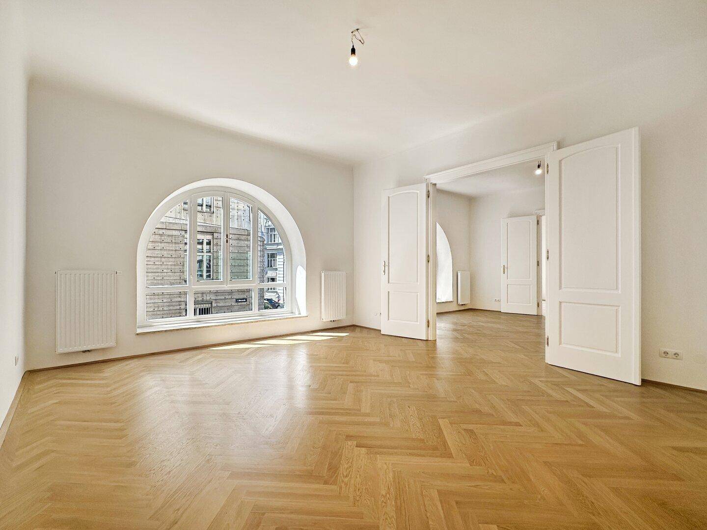Heller Raum mit Fischgrätparkett und einem großen Rundbogenfenster, das den Blick auf die umliegenden Gebäude freigibt.