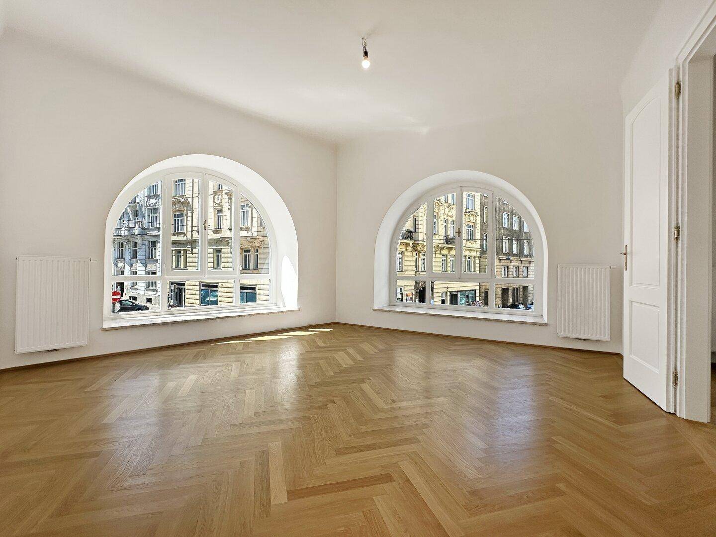 Helles Zimmer mit zwei großen Rundbogenfenstern, die viel Tageslicht hereinlassen und den Blick auf die Stadt bieten.