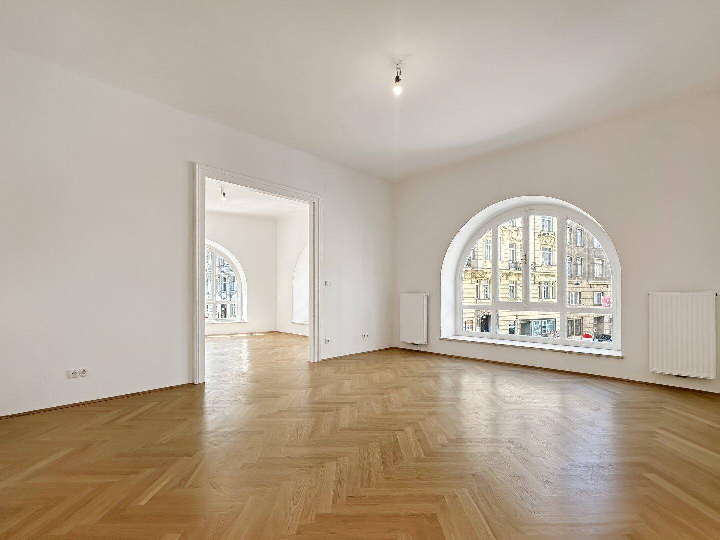 Geräumiger Wohnbereich mit glänzendem Fischgrätparkett und einem großen Rundbogenfenster mit Stadtblick.
