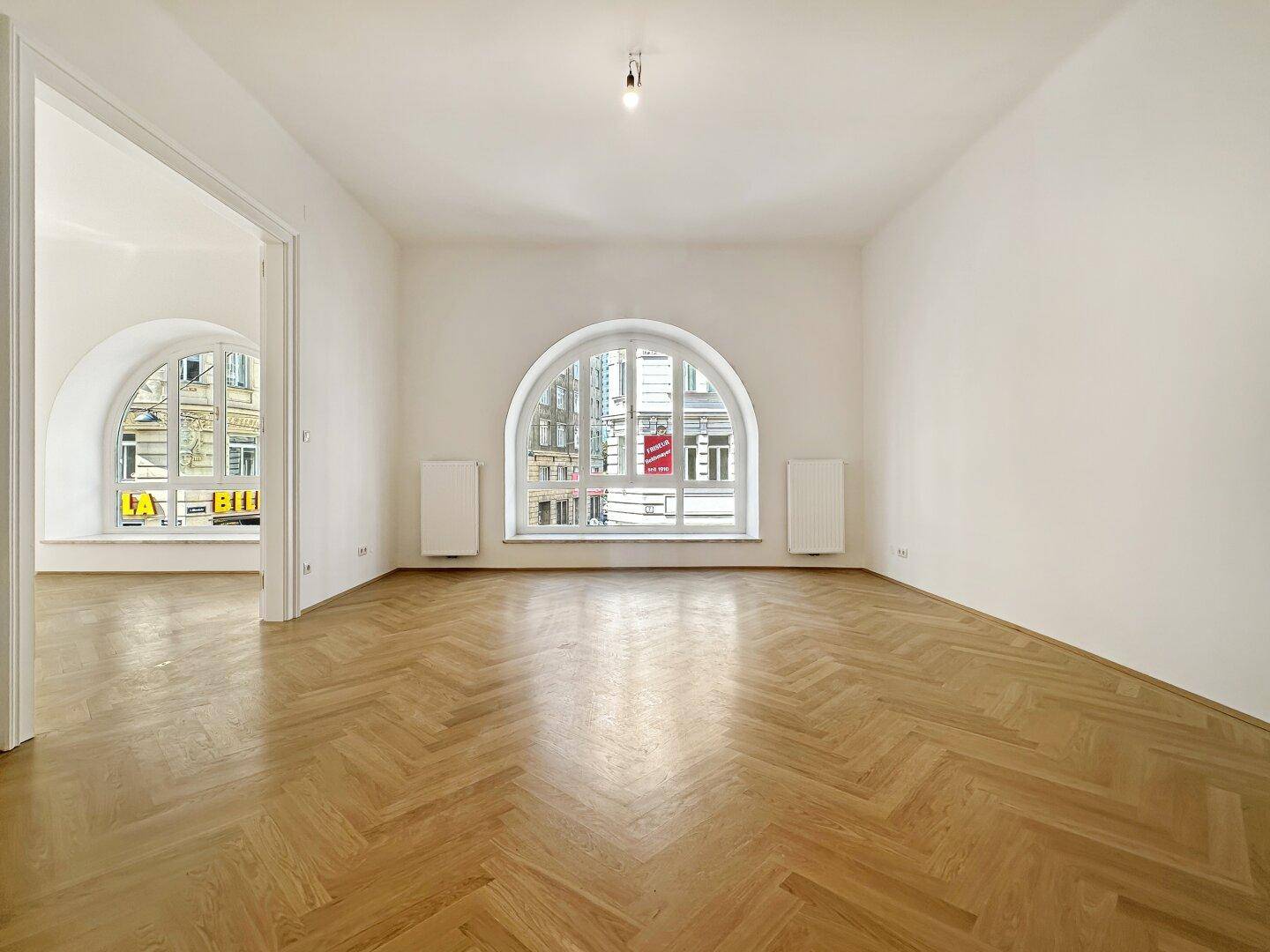 Großes, helles Zimmer mit elegantem Fischgrätparkett und einem markanten Rundbogenfenster zur Straße.