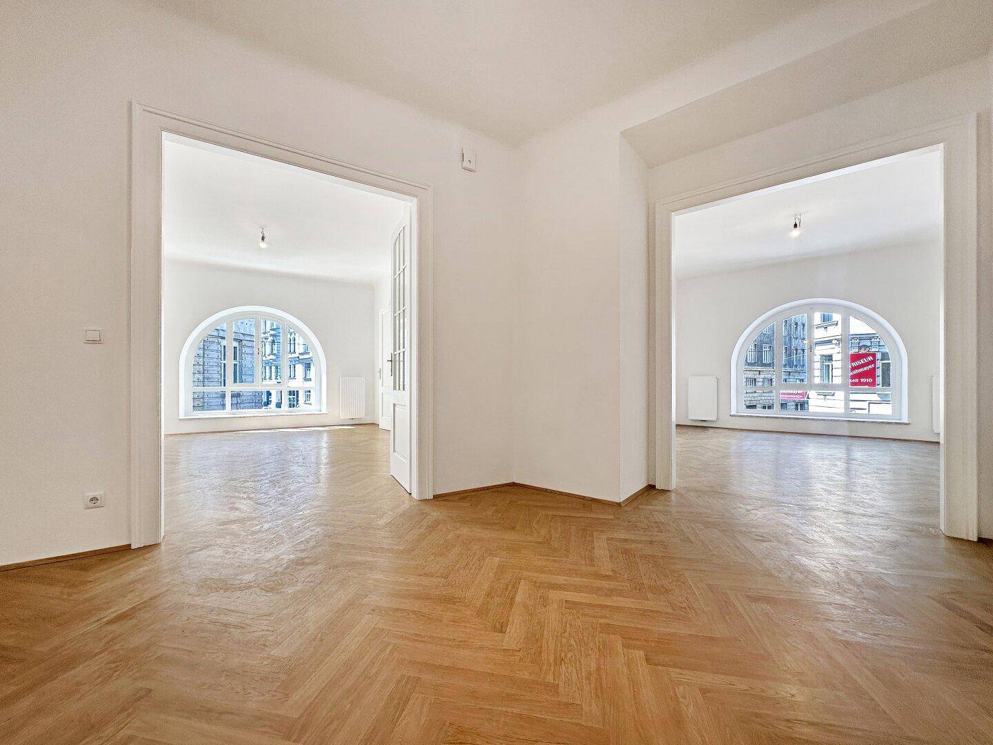 Heller Raum mit Fischgrätparkett und zwei großen Rundbogenfenstern, die den Blick auf die Stadt freigeben.