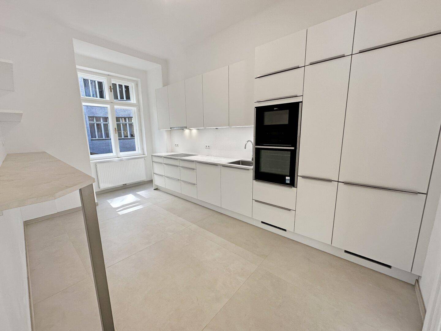 Moderne weiße Einbauküche mit integrierten Geräten und einem Fenster, das den Raum erhellt.