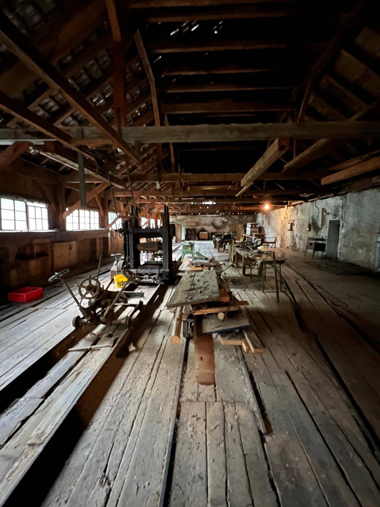 Geräumiger Werkstattraum mit Holzboden, alten Maschinen und sichtbaren Holzbalken der Dachkonstruktion.