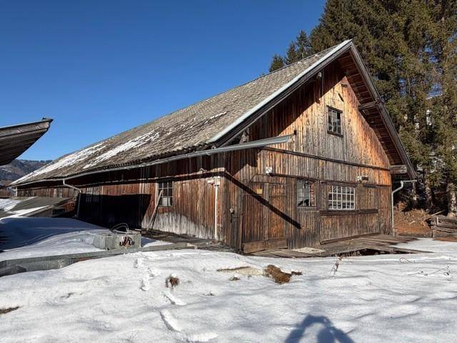 Lange Seite eines rustikalen Holzgebäudes mit verschneitem Boden und Blick auf bewaldete Hügel.
