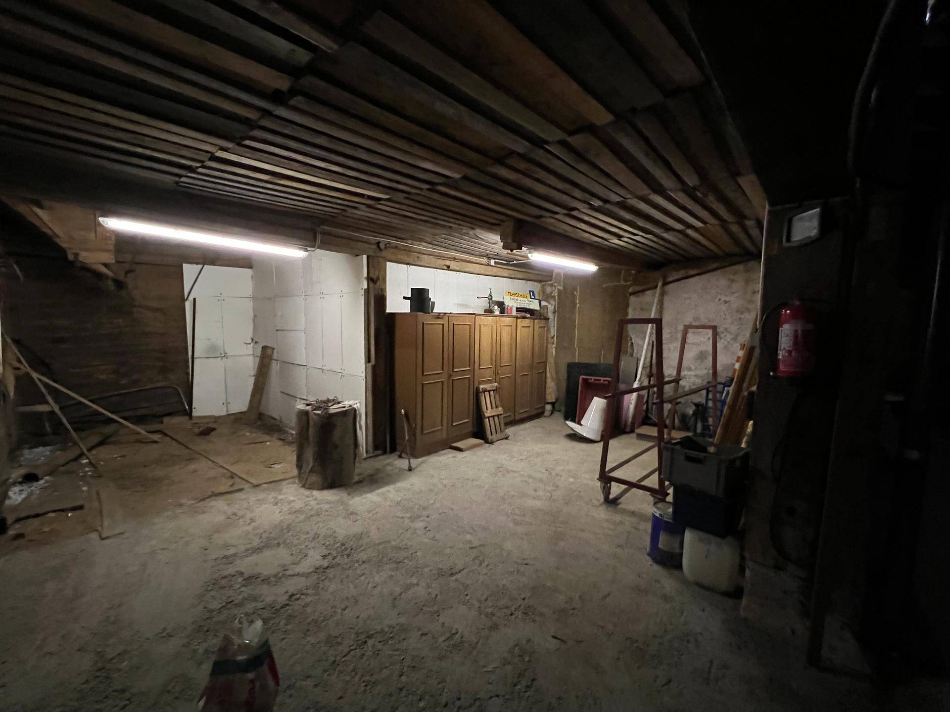 Dunkler Lagerraum mit Holzbalkendecke, Betonboden und verschiedenen gelagerten Gegenständen.