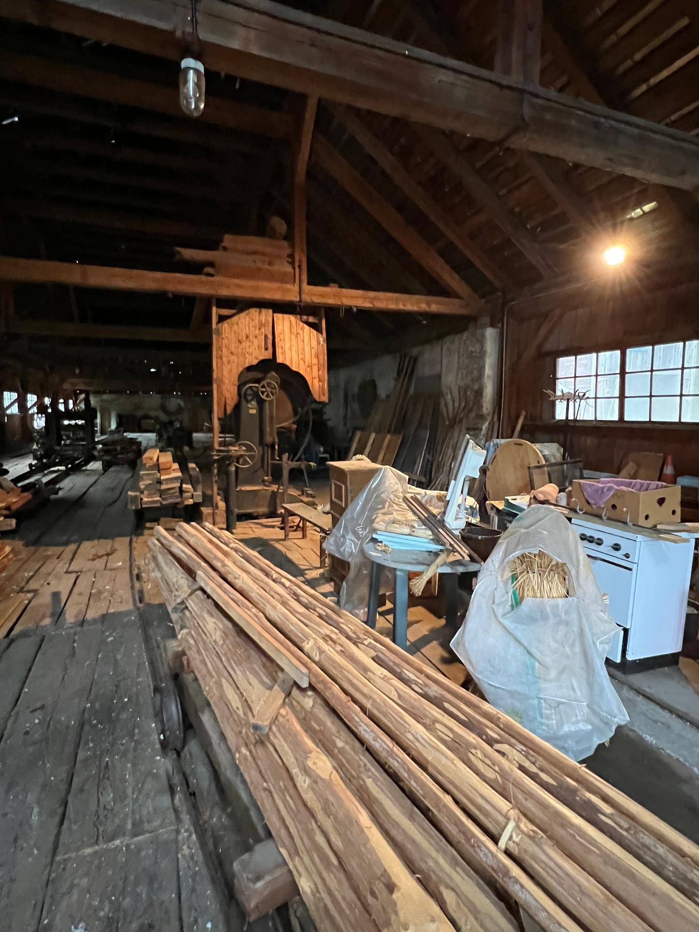 Werkstattraum mit Holzboden, gelagertem Holz und verschiedenen Geräten unter sichtbaren Dachbalken.