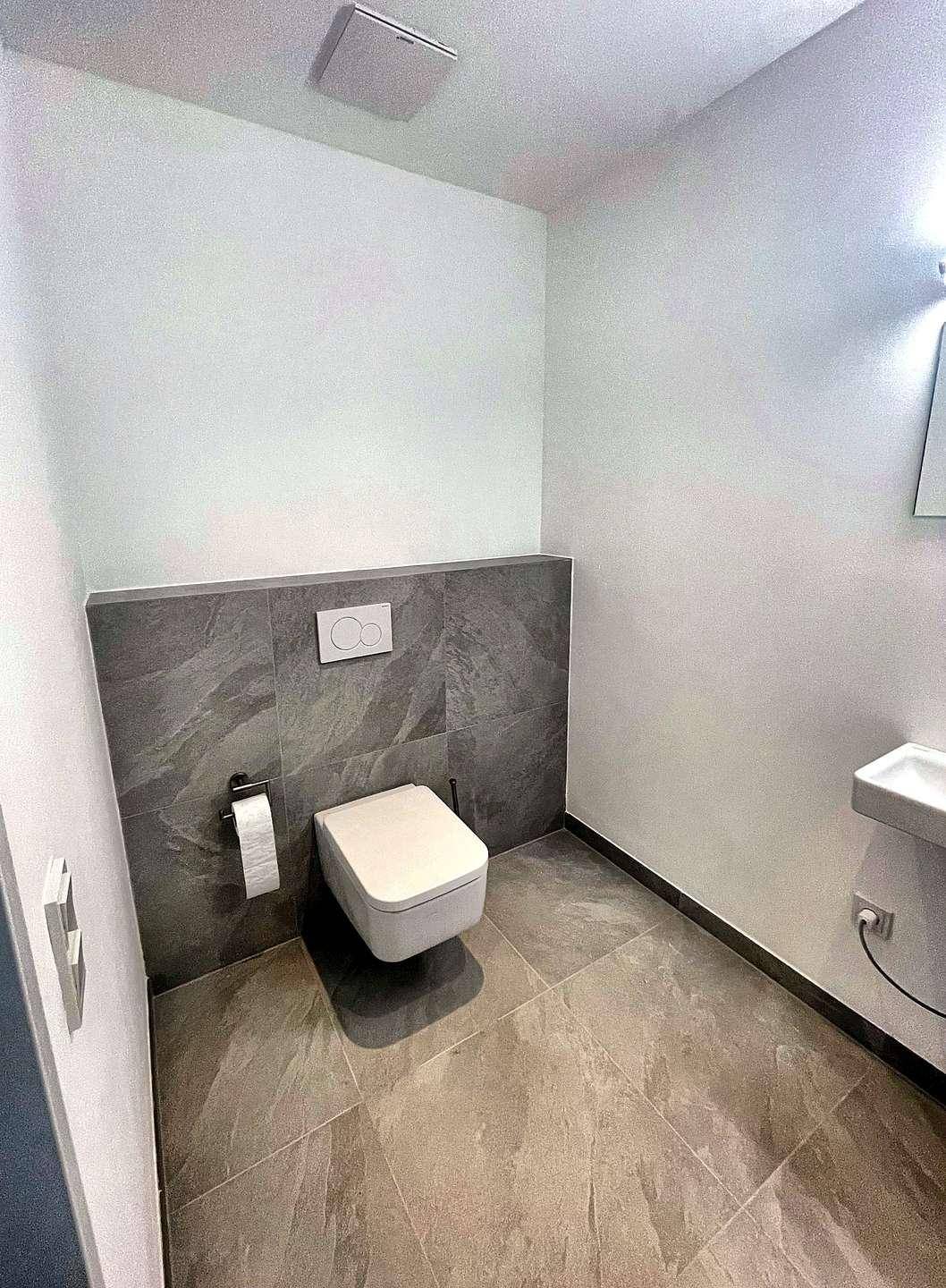 Helles Gäste-WC mit wandhängender Toilette, kleinem Waschbecken und grauen Wandfliesen.