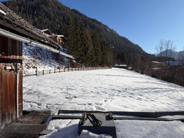 Verschneites Feld neben einem Holzgebäude mit bewaldeten Bergen und blauem Himmel im Hintergrund.