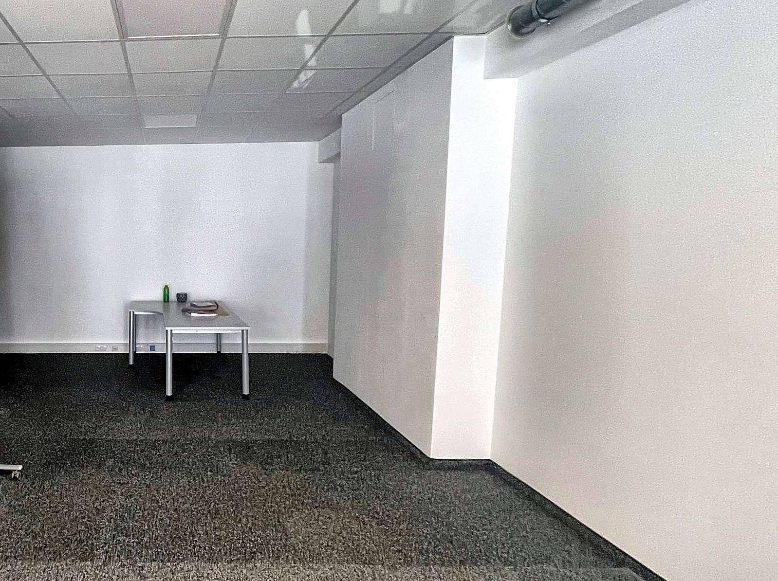 Heller Bürobereich mit Teppichboden, weißen Wänden und einem kleinen Tisch in der Ecke.