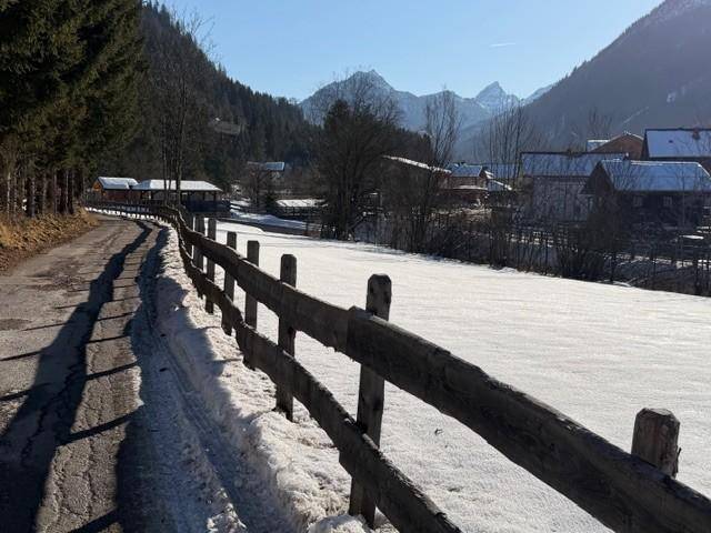 Verschneite Straße und Feld mit Holzzaun, die zu einem Dorf und Bergen in der Ferne führen.