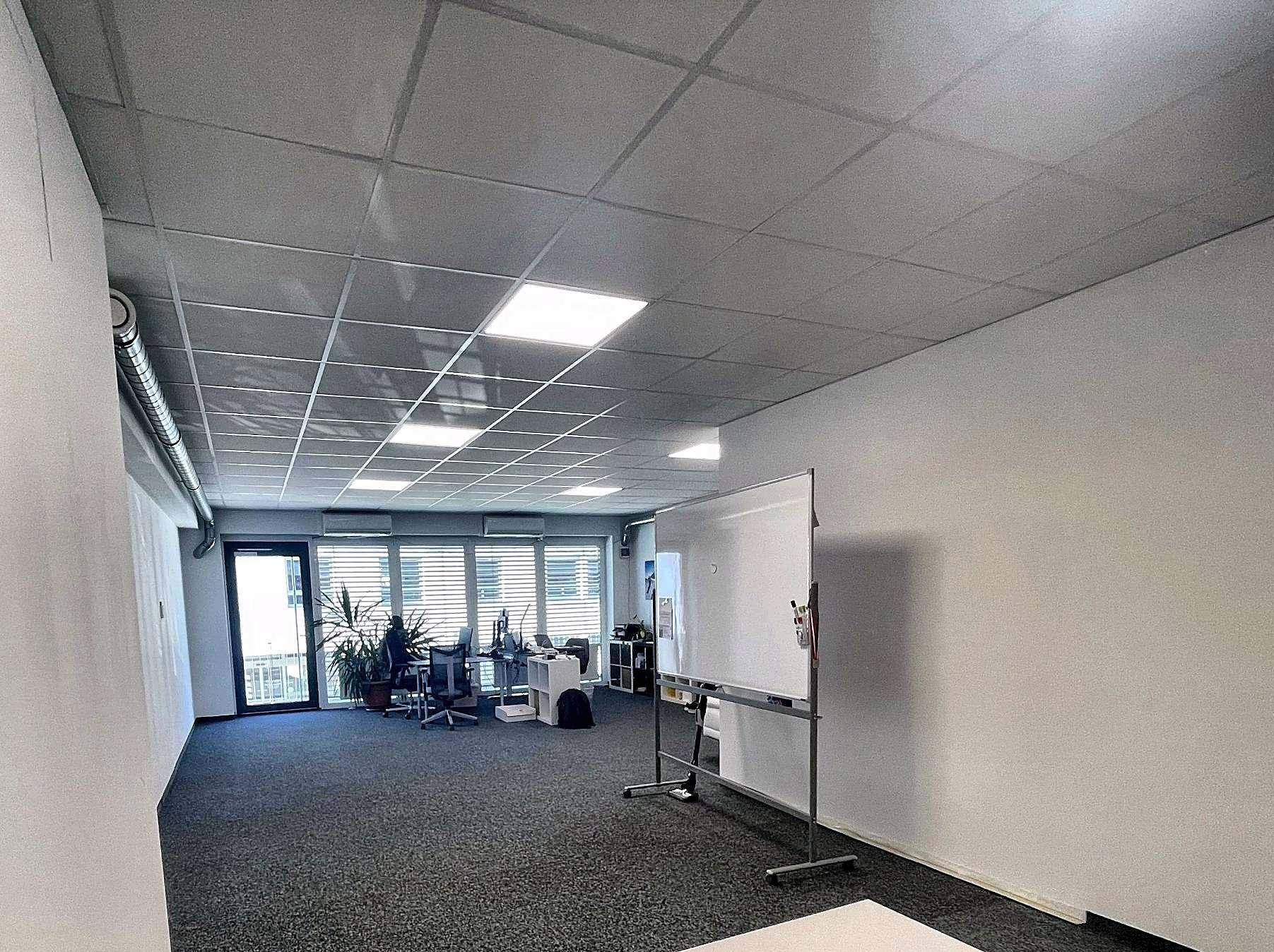 Weitläufiges Büro mit Teppichboden, abgehängter Decke und einer großen Whiteboard-Tafel.