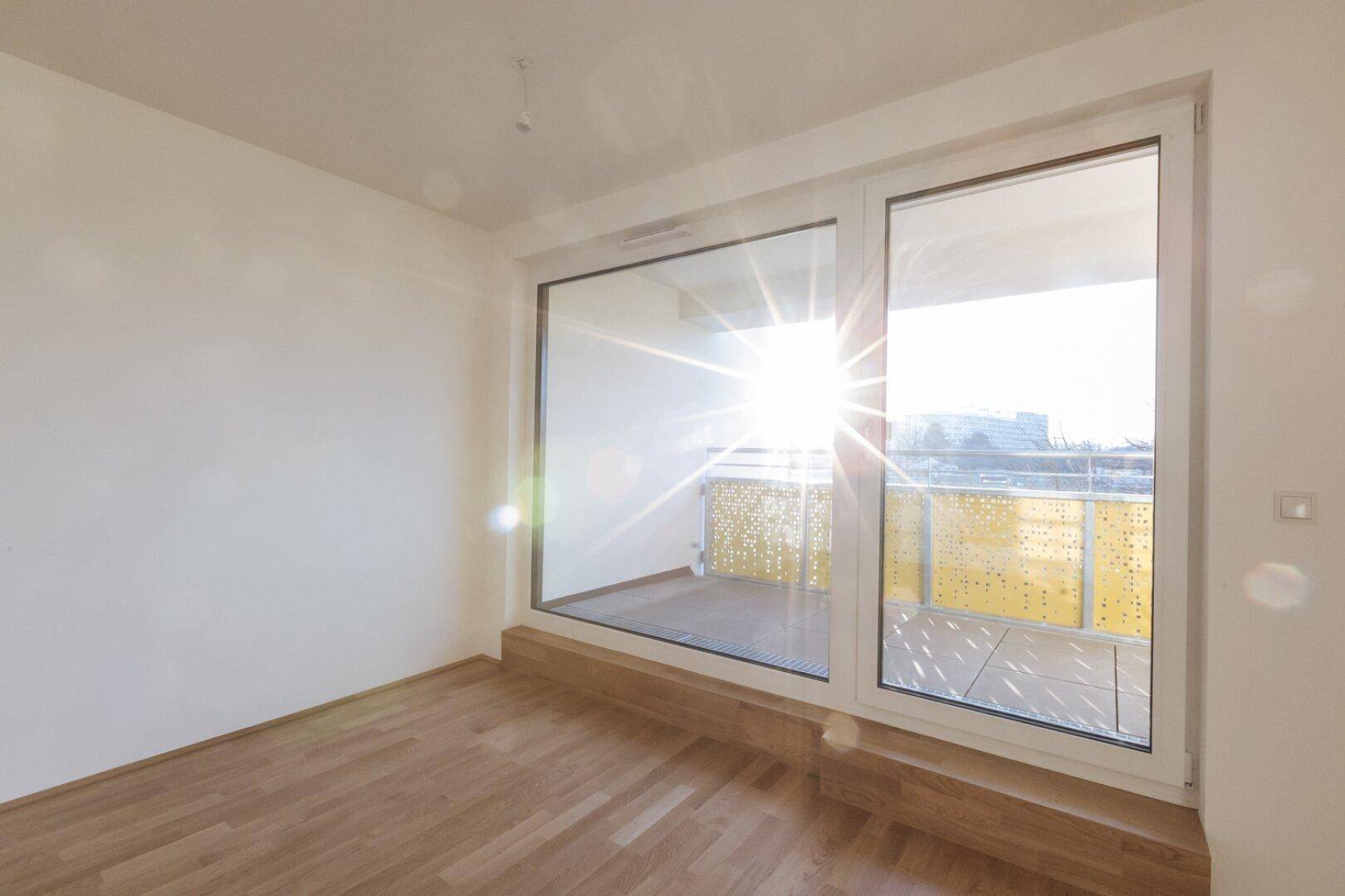 Lichtdurchfluteter Raum mit Parkettboden und breiter Fensterfront zum sonnigen Balkon.