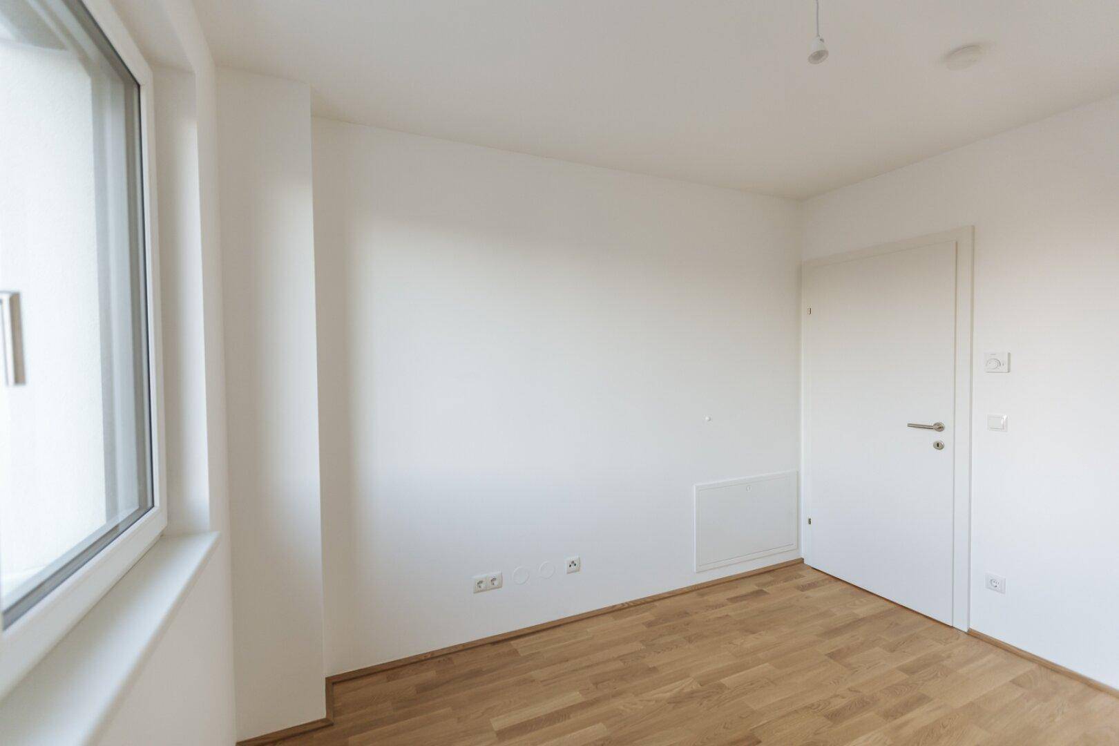 Helles Schlafzimmer mit Parkettboden und einem Fenster, das viel Tageslicht hereinlässt.
