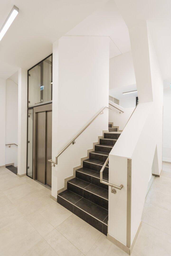 Treppenhaus und Aufzug in einem modernen Gebäude, mit klaren Linien und hellen Materialien.