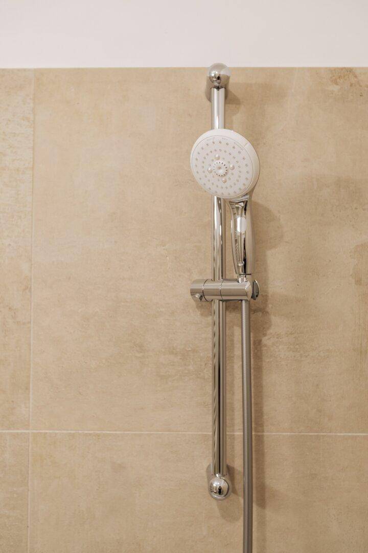 Nahaufnahme einer modernen Duschstange mit Handbrause an einer beigefliesten Wand.