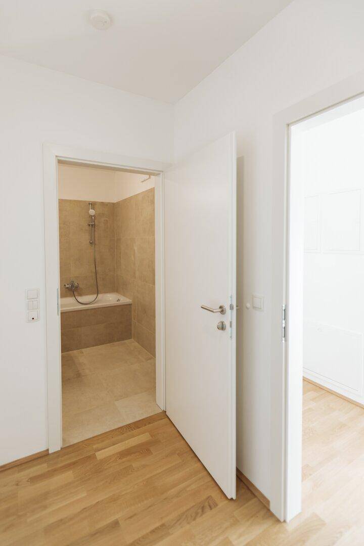 Heller Flur mit Holzboden, der zu einem Badezimmer mit Badewanne führt.