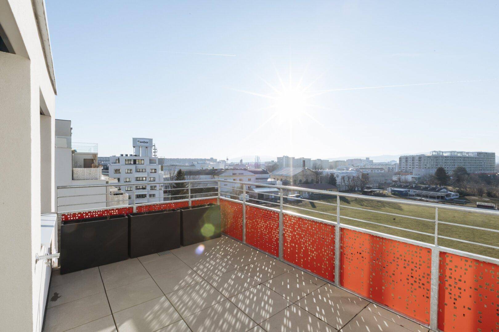 Sonnige Terrasse mit modernen orangefarbenen Geländern und weitem Blick über die Stadt.